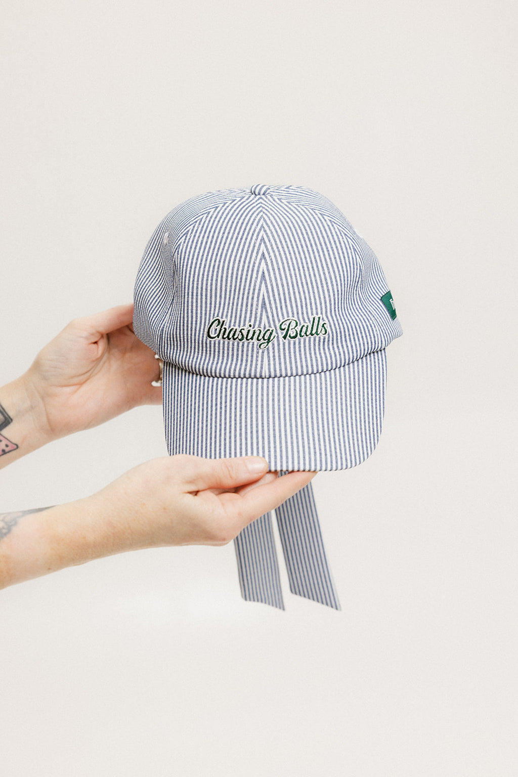 A person holding a blue and white seersucker baseball cap with the text 'Chasing Balls' embroidered on the front and an oversized bow at the back.