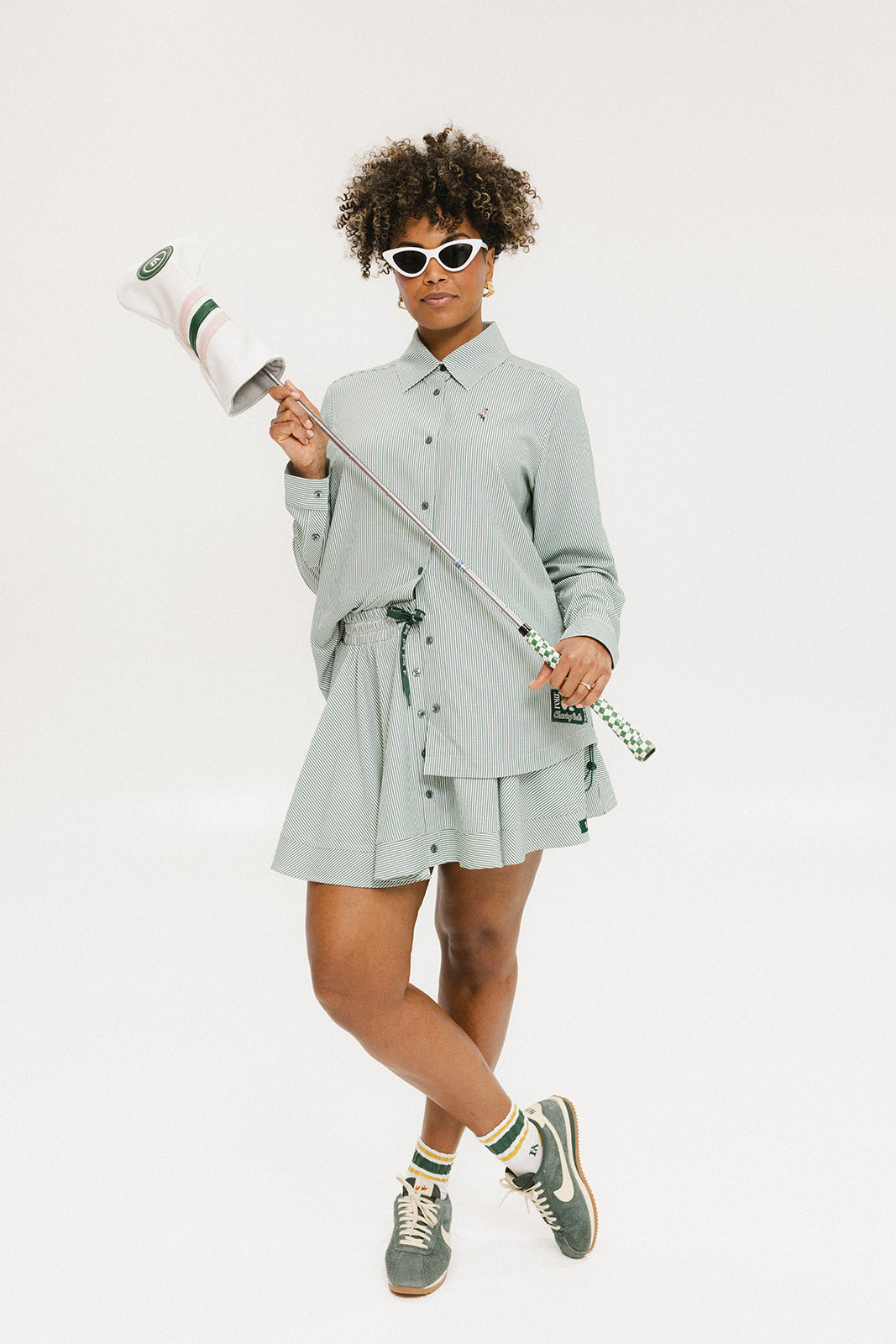 A woman wearing a green striped button-up shirt with a collar, sleeve cuffs, and side seam hem, paired with a matching skirt, sunglasses, and green shoes.