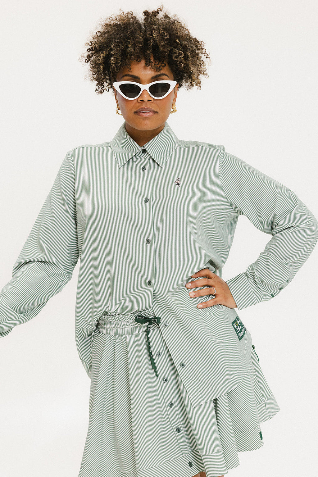 A woman wearing a green striped button-up shirt with a collar, sleeve cuffs, and side seam hem, paired with a matching skirt, sunglasses, and green shoes.