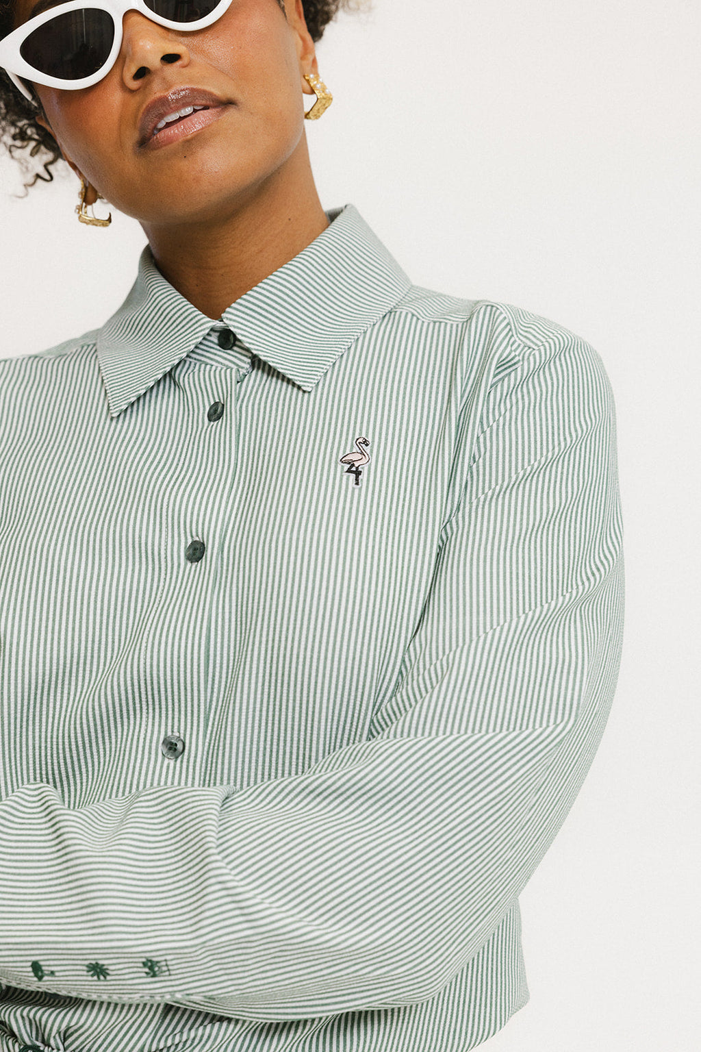 A woman wearing a green striped button-up shirt with a collar, sleeve cuffs, and side seam hem, paired with a matching skirt, sunglasses, and green shoes.
