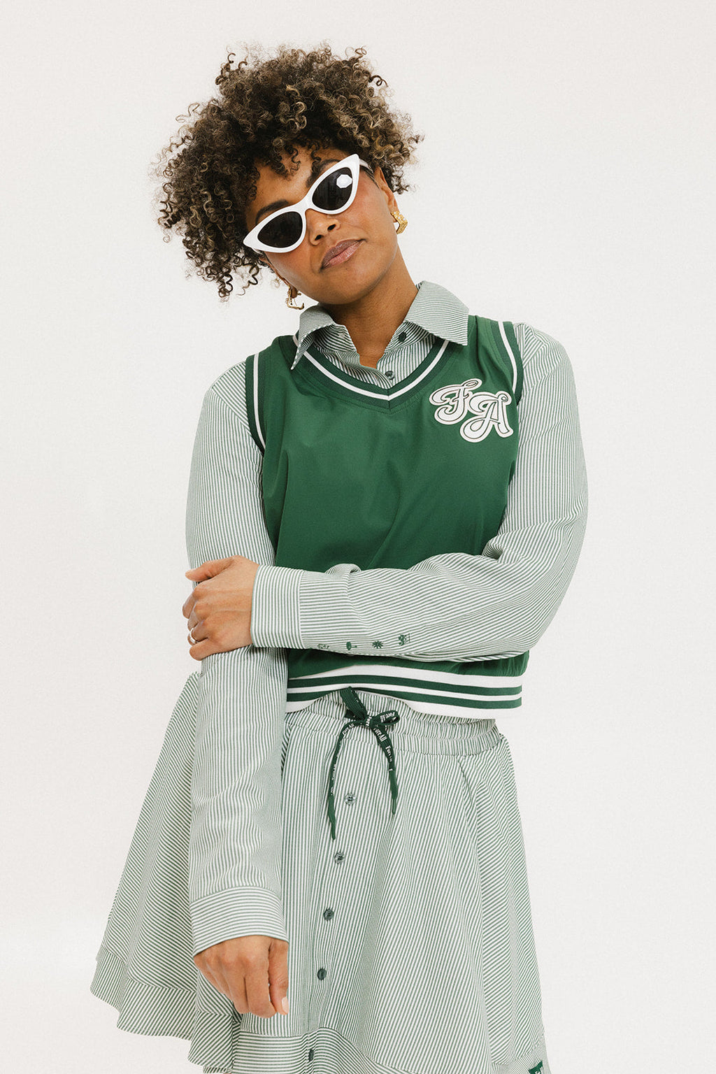 A woman wearing a green striped button-up shirt with a collar, sleeve cuffs, and side seam hem, paired with a matching skirt, sunglasses, and green shoes.