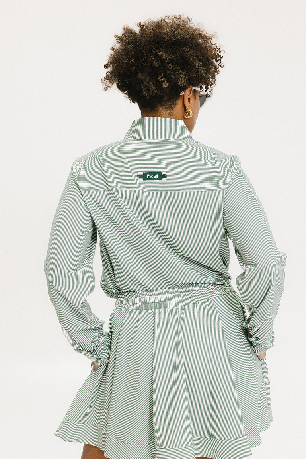 A woman wearing a green striped button-up shirt with a collar, sleeve cuffs, and side seam hem, paired with a matching skirt, sunglasses, and white shoes.