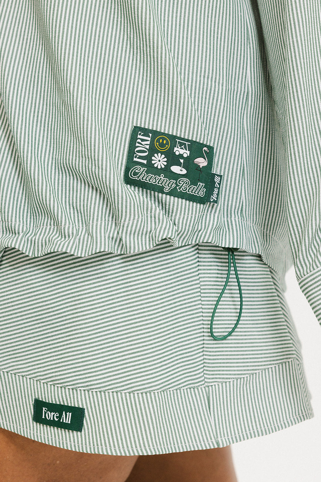 A woman wearing a green striped button-up shirt with a collar, sleeve cuffs, and side seam hem, paired with a matching skirt, sunglasses, and green shoes.