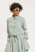 A woman wearing a green striped button-up shirt with a collar, sleeve cuffs, and side seam hem, paired with a matching skirt, sunglasses, and white shoes.