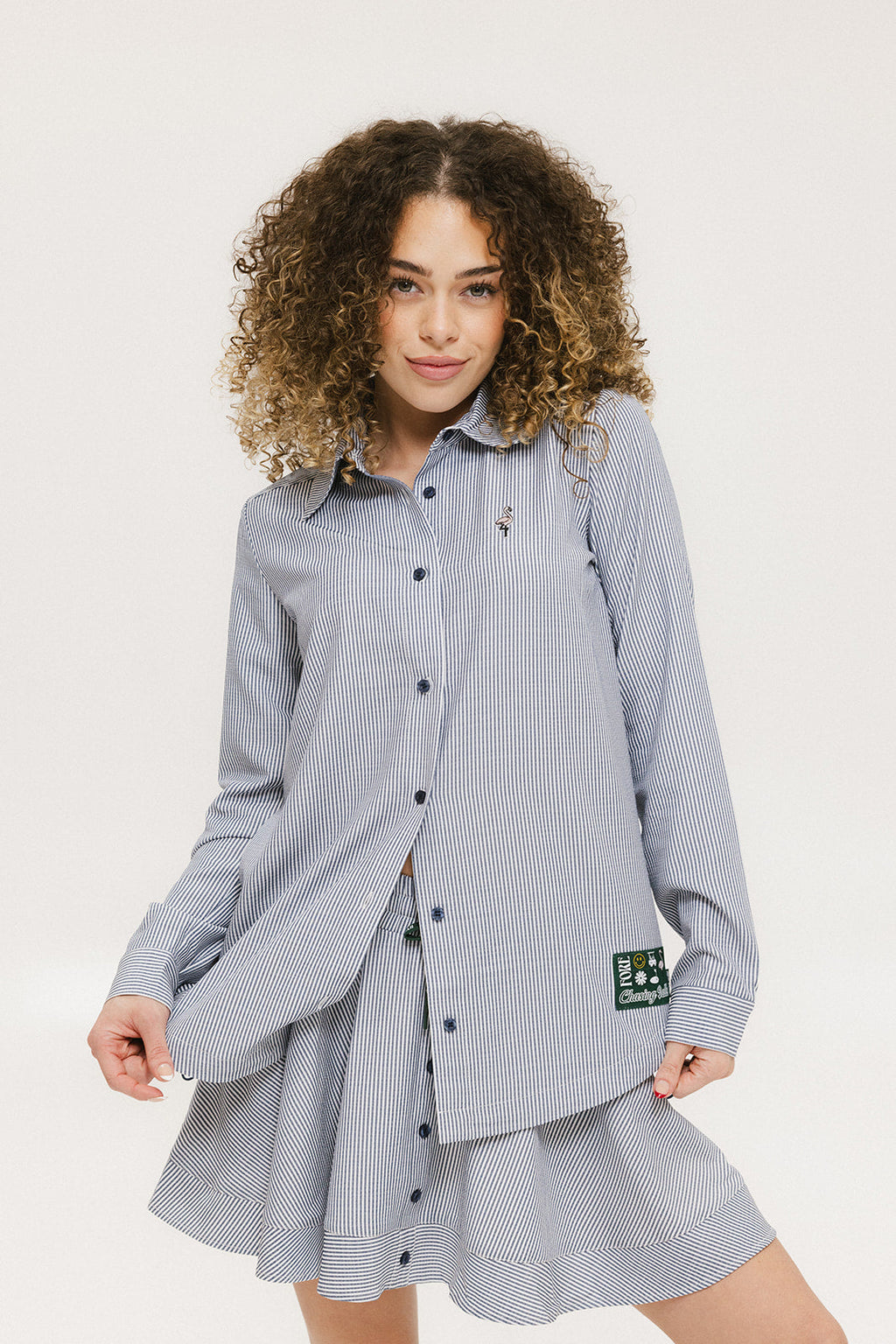 A woman wearing a long, button-up shirt with indigo stripes, featuring a collar, button-down front, and elastic toggles at the side seam hem and sleeve cuffs.