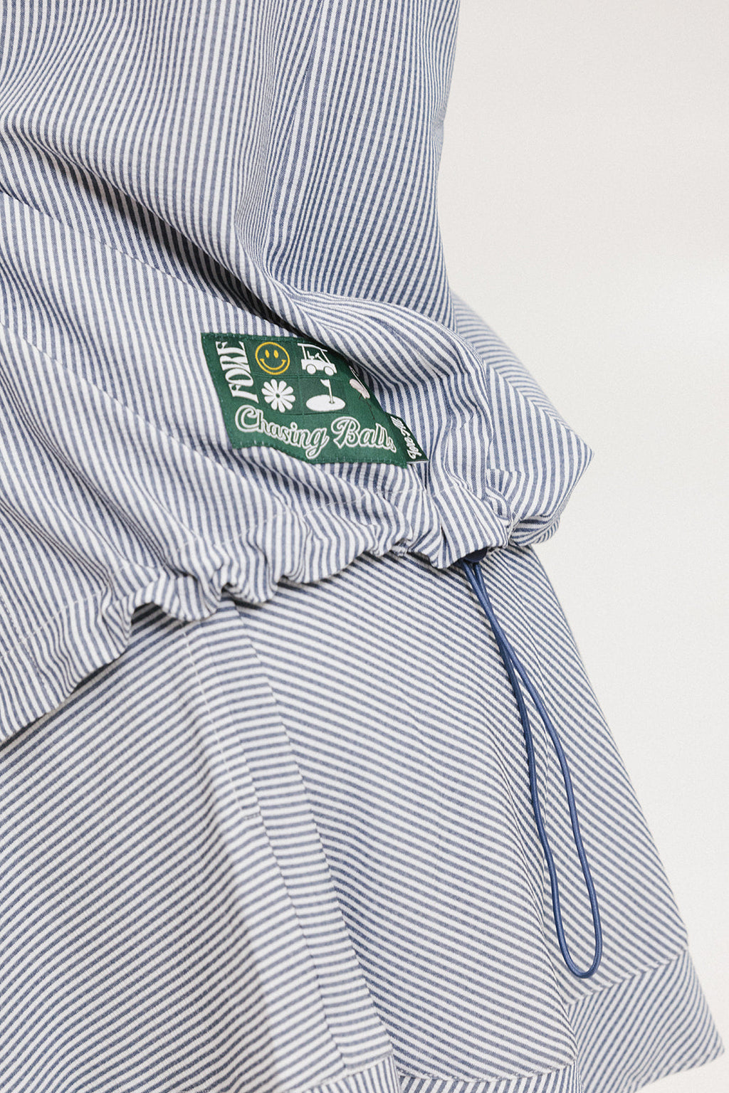 A woman wearing a long, button-up shirt with indigo stripes, featuring a collar, button-down front, and elastic toggles at the side seam hem and sleeve cuffs
