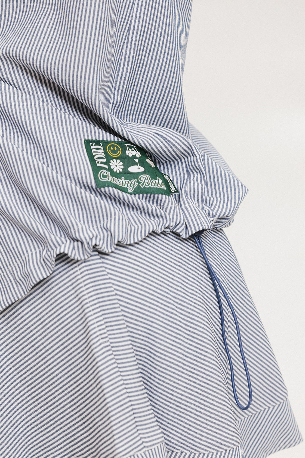 A woman wearing a long, button-up shirt with indigo stripes, featuring a collar, button-down front, and elastic toggles at the side seam hem and sleeve cuffs