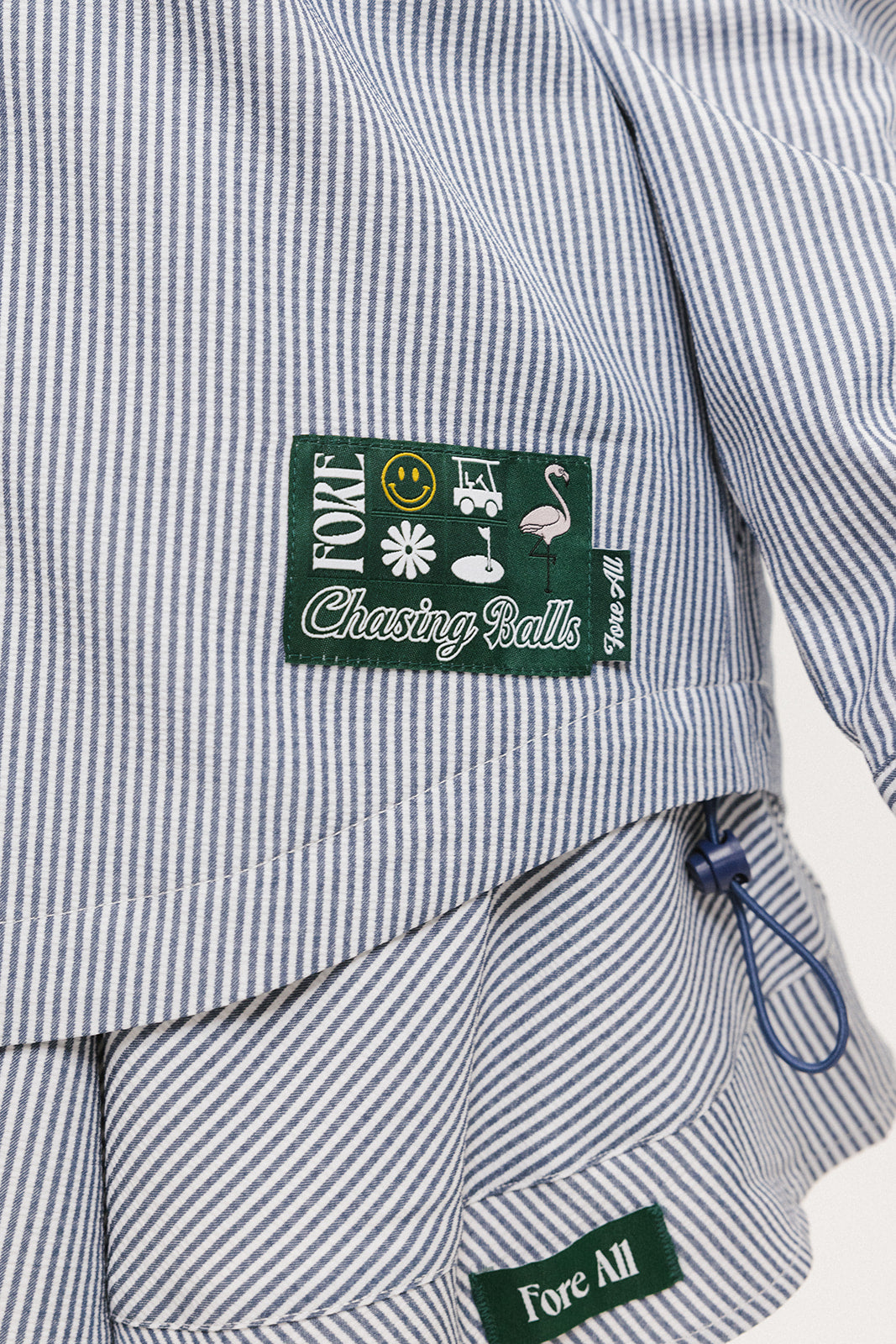 A woman wearing a long, button-up shirt with indigo stripes, featuring a collar, button-down front, and elastic toggles at the side seam hem and sleeve cuffs