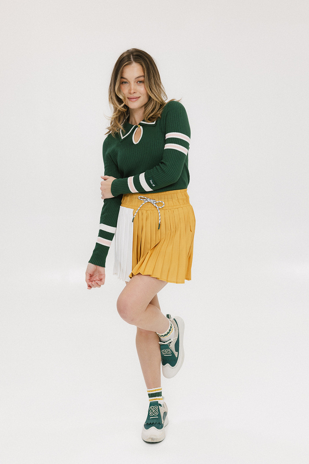 A woman wearing a green ribbed keyhole polo with white stripe accents on the sleeves and a gold smiley face button at the center front neck, paired with a yellow and white skirt.