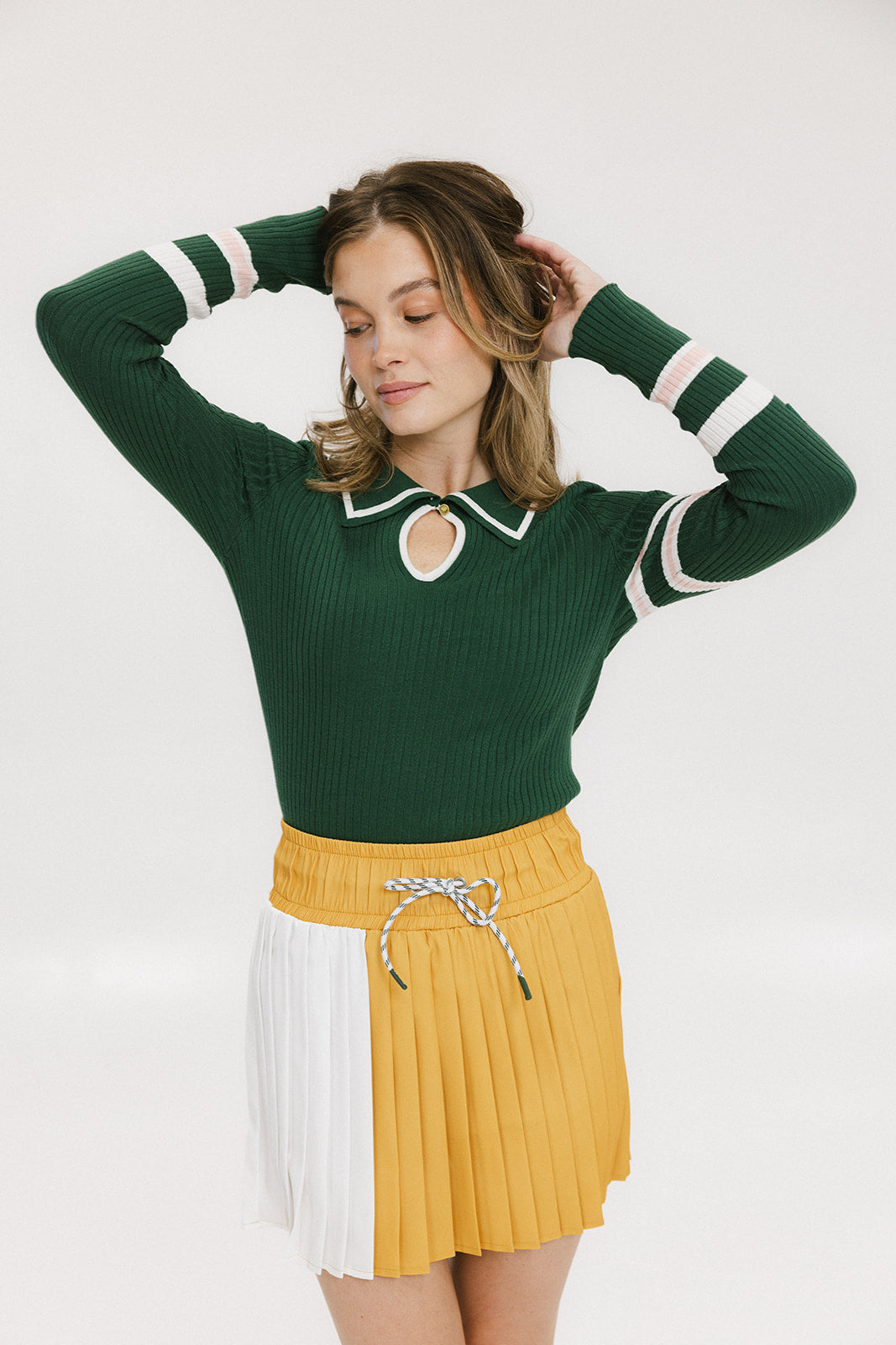 A woman wearing a green ribbed keyhole polo with white stripe accents on the sleeves and a gold smiley face button at the center front neck, paired with a yellow and white skirt.