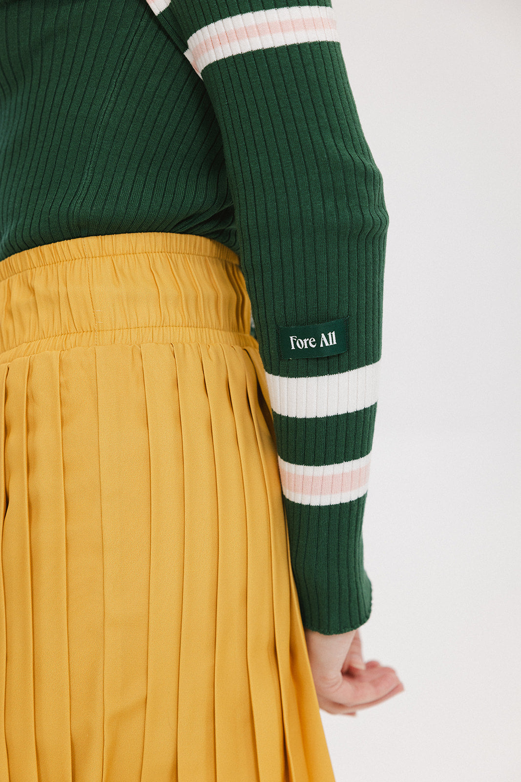 A woman wearing a green ribbed keyhole polo with white stripe accents on the sleeves and a gold smiley face button at the center front neck, paired with a yellow and white skirt.