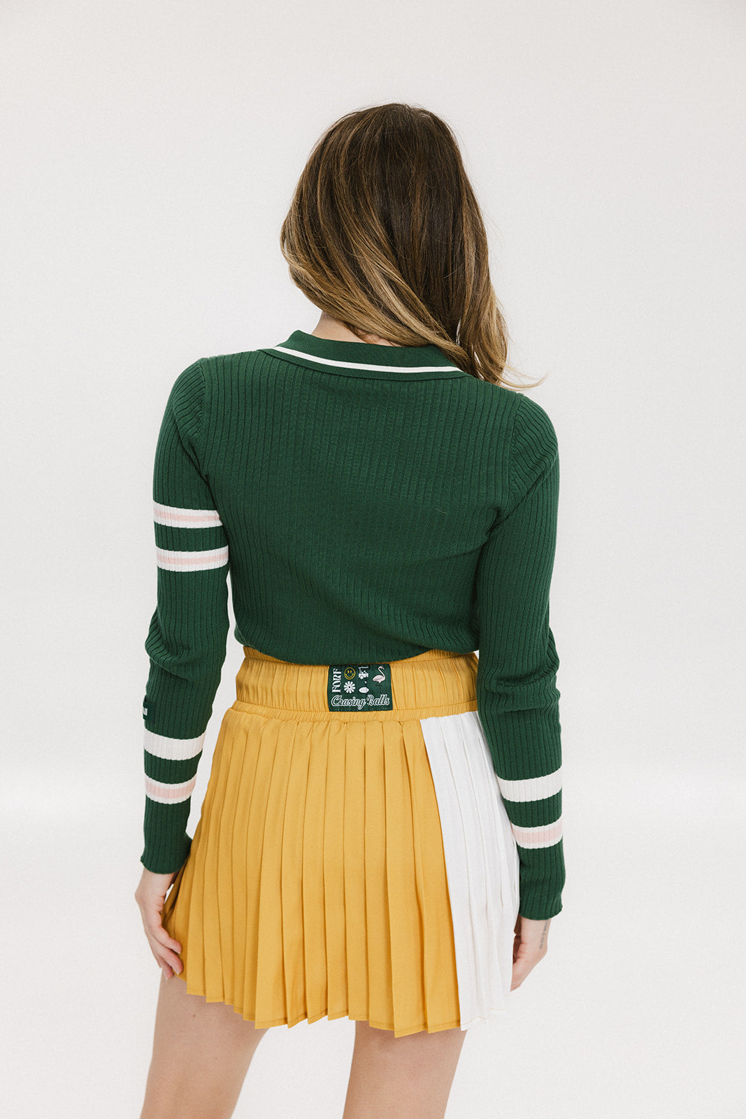 A woman wearing a green ribbed keyhole polo with white stripe accents on the sleeves and a gold smiley face button at the center front neck, paired with a yellow and white skirt.