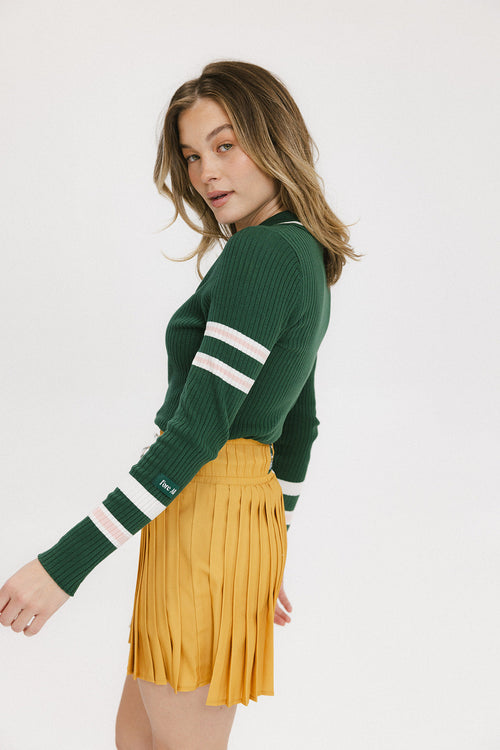 A woman wearing a green ribbed keyhole polo with white stripe accents on the sleeves and a gold smiley face button at the center front neck, paired with a yellow and white skirt.