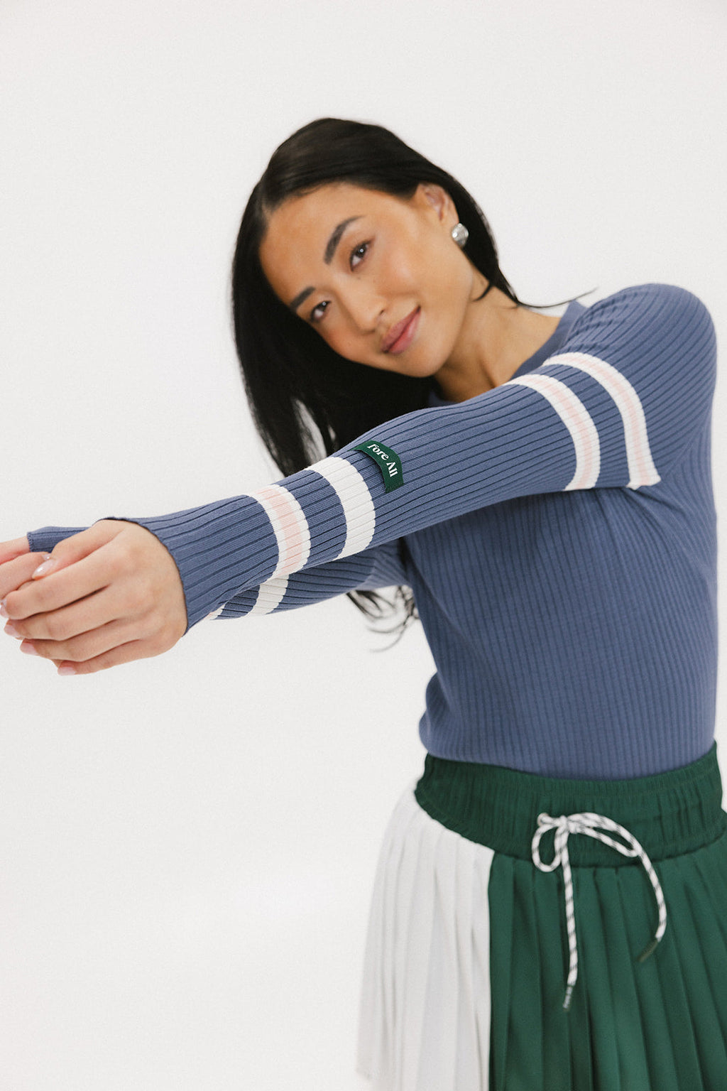A woman wearing a blue ribbed keyhole polo with white stripe accents on the sleeves and a green and white pleated skirt.