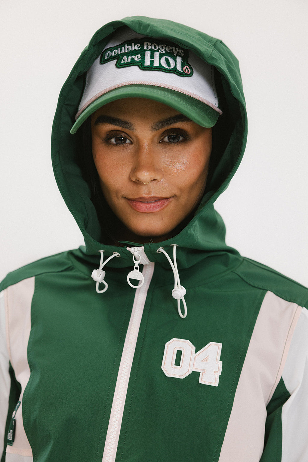 A woman wearing a long-sleeved green and pink warm-up jacket with a hood, zipper pockets, and colorblocking. She also sports a matching green cap and appears to be standing against a white background.