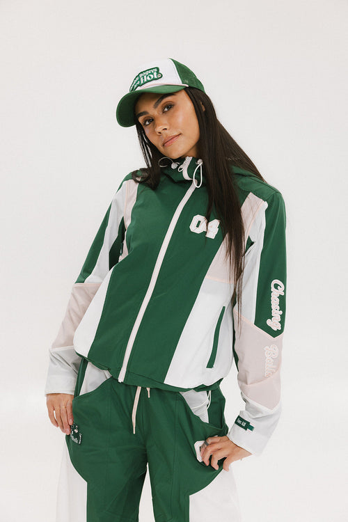 A woman wearing a long-sleeved green and pink warm-up jacket with a hood, zipper pockets, and colorblocking. She also sports a matching green cap and appears to be standing against a white background.