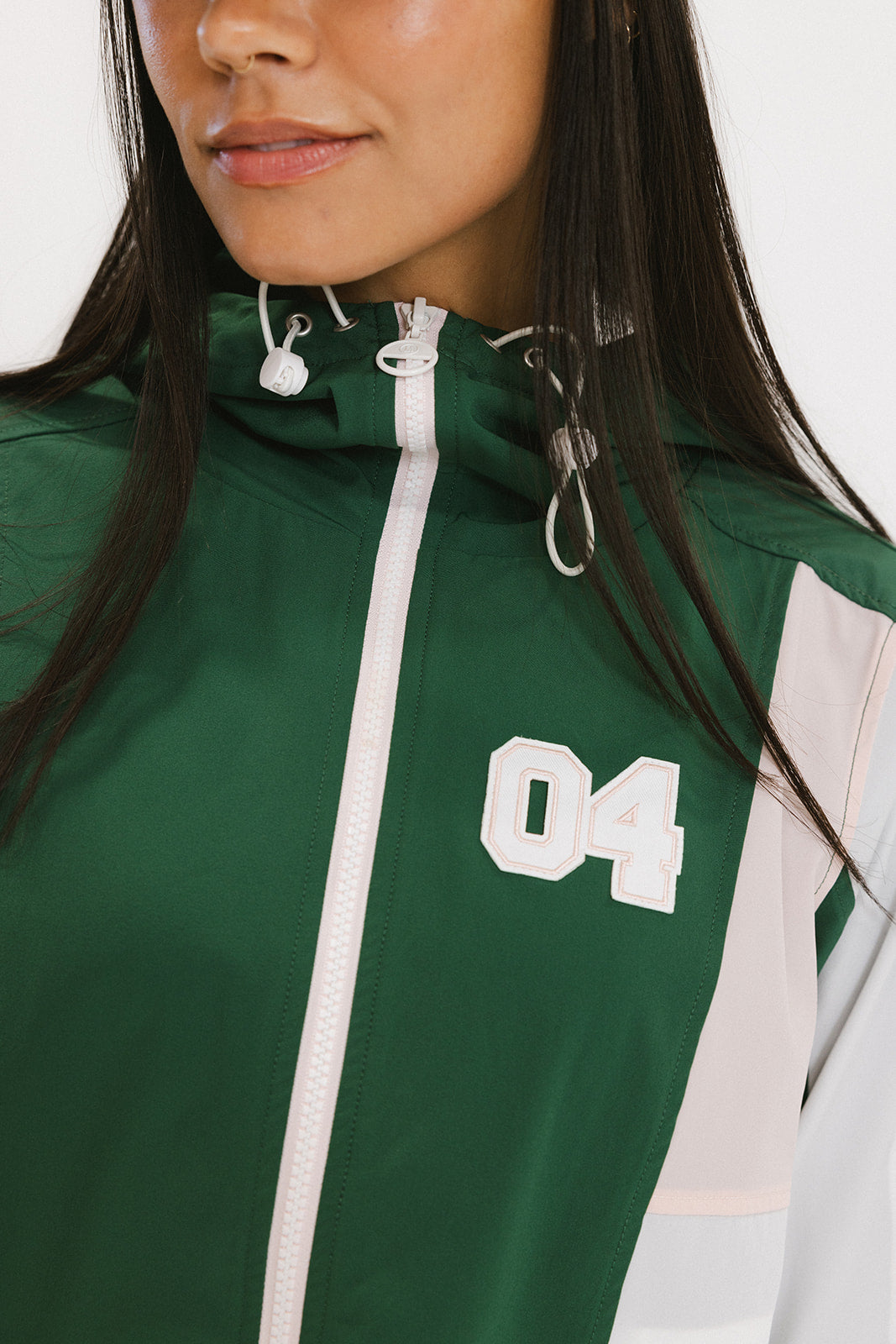 A woman wearing a long-sleeved green and pink warm-up jacket with a hood, zipper pockets, and colorblocking. She also sports a matching green cap and appears to be standing against a white background.