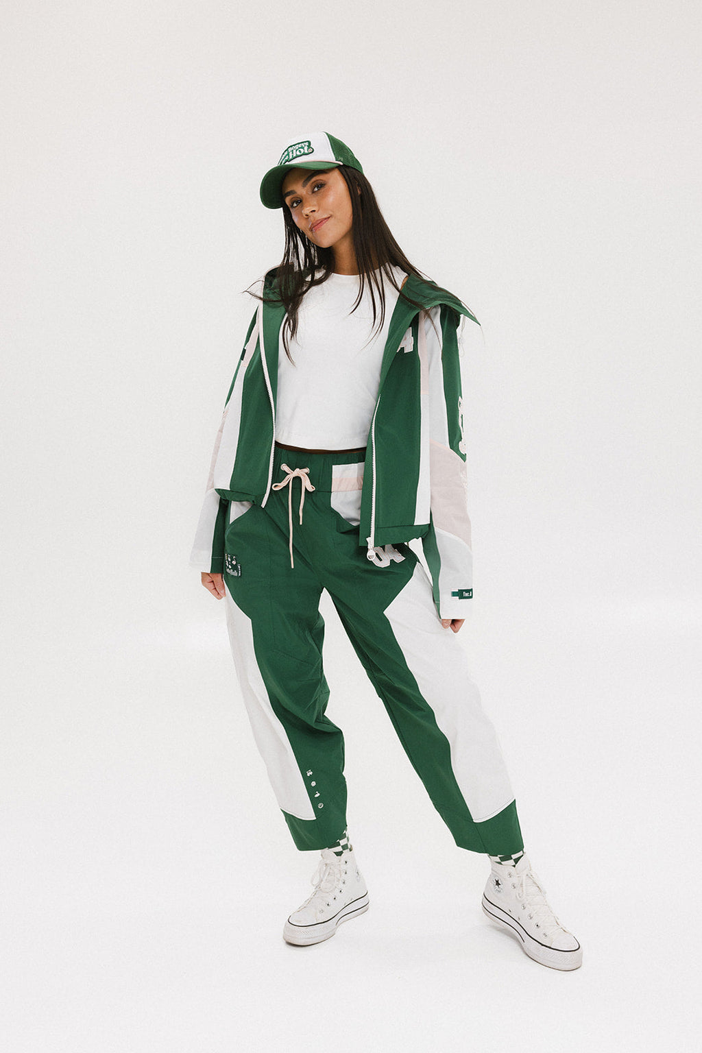 A woman wearing a long-sleeved green and pink warm-up jacket with a hood, zipper pockets, and colorblocking. She also sports a matching green cap and appears to be standing against a white background.