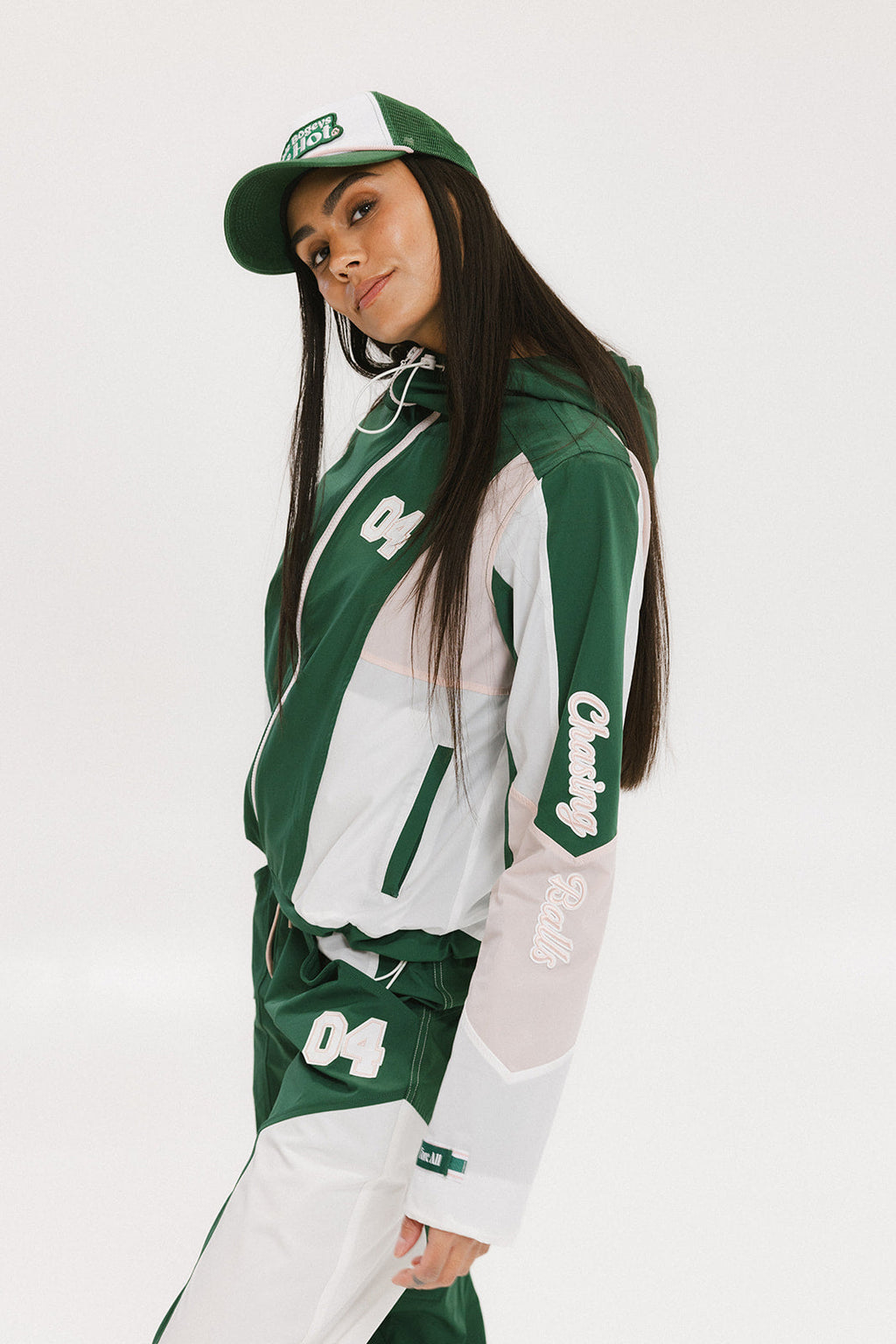 A woman wearing a long-sleeved green and pink warm-up jacket with a hood, zipper pockets, and colorblocking. She also sports a matching green cap and appears to be standing against a white background.