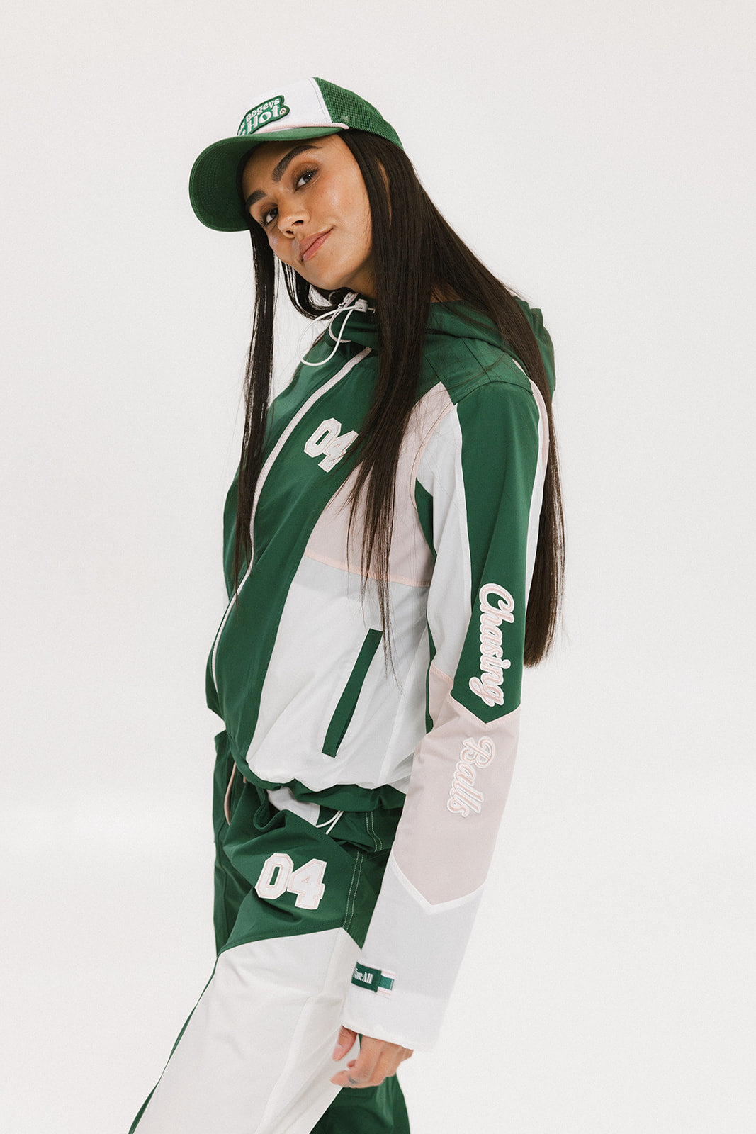 A woman wearing a long-sleeved green and pink warm-up jacket with a hood, zipper pockets, and colorblocking. She also sports a matching green cap and appears to be standing against a white background.