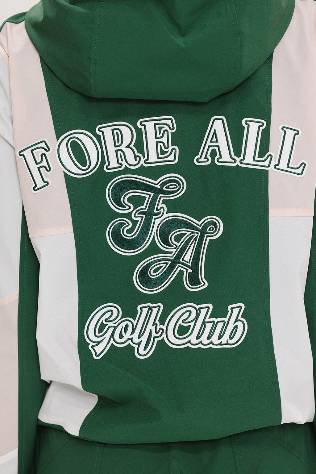 A woman wearing a long-sleeved green and pink warm-up jacket with a hood, zipper pockets, and colorblocking. She also sports a matching green cap and appears to be standing against a white background.