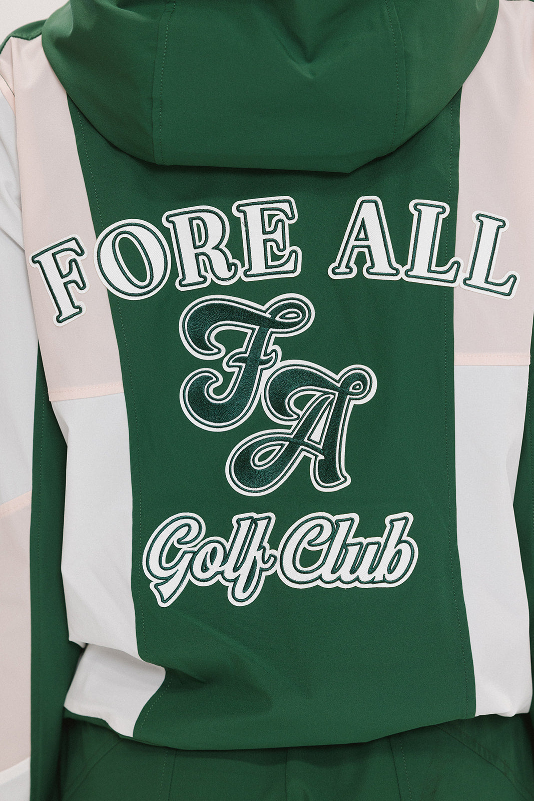 A woman wearing a long-sleeved green and pink warm-up jacket with a hood, zipper pockets, and colorblocking. She also sports a matching green cap and appears to be standing against a white background.