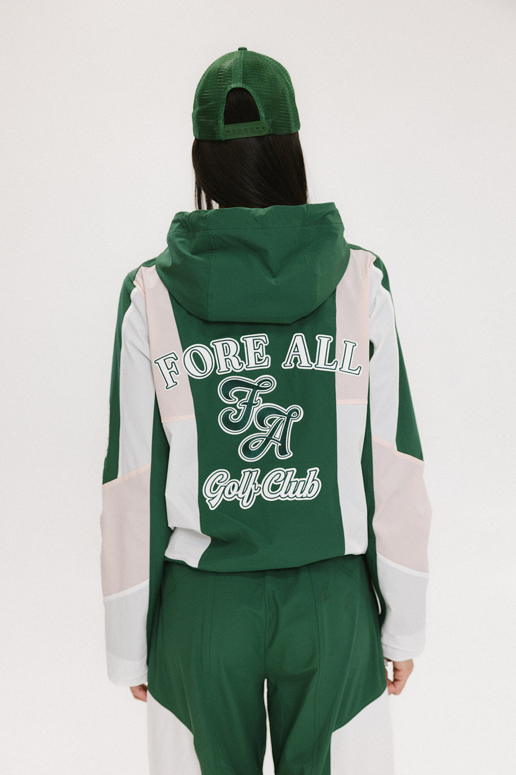 A woman wearing a long-sleeved green and pink warm-up jacket with a hood, zipper pockets, and colorblocking. She also sports a matching green cap and appears to be standing against a white background.