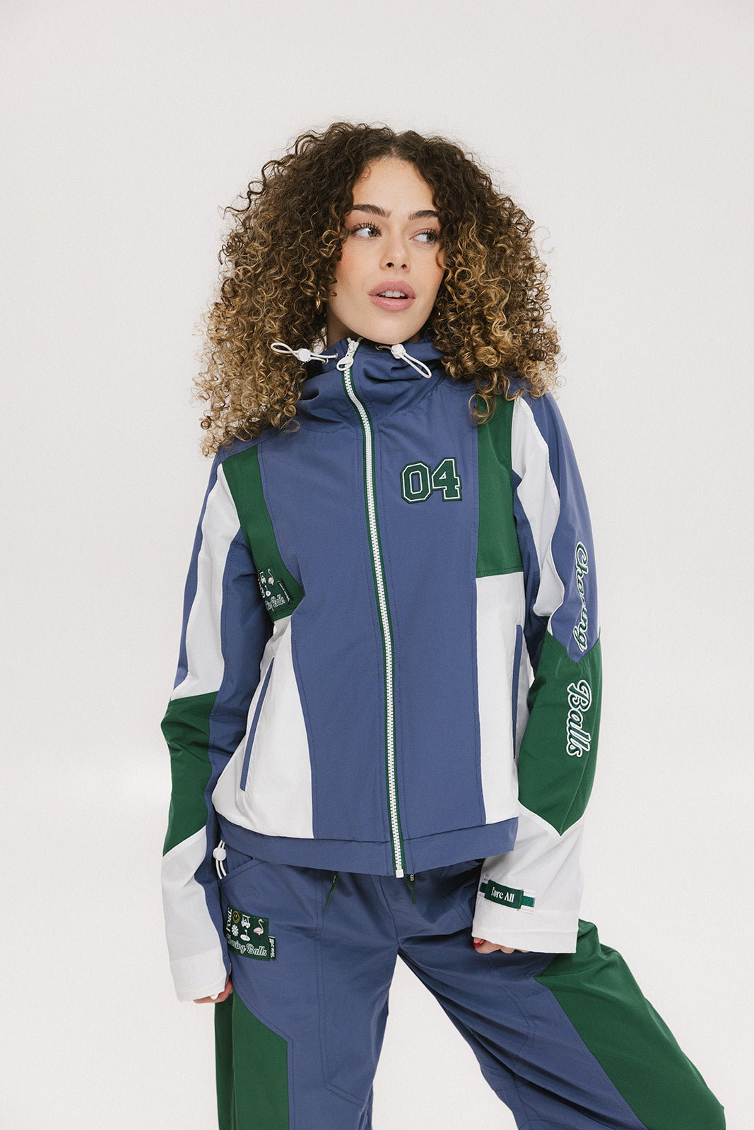 A female model wearing a blue and green color-blocked warm-up jacket with zipper pockets and a high neck collar.