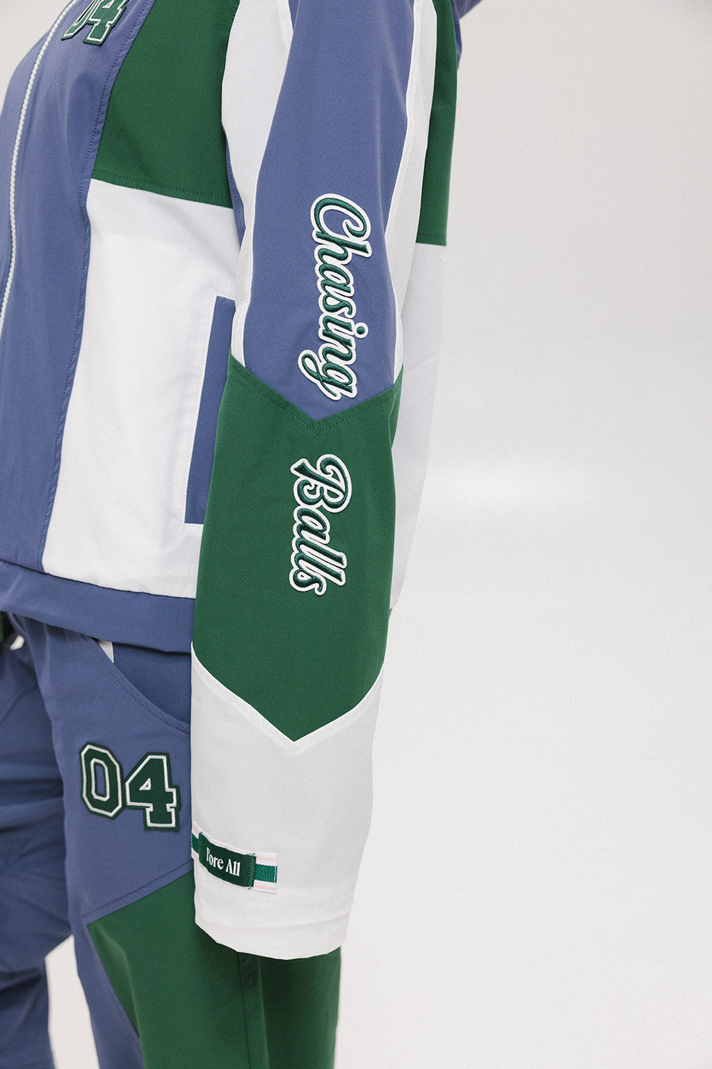 A female model wearing a blue and green color-blocked warm-up jacket with zipper pockets and a high neck collar.