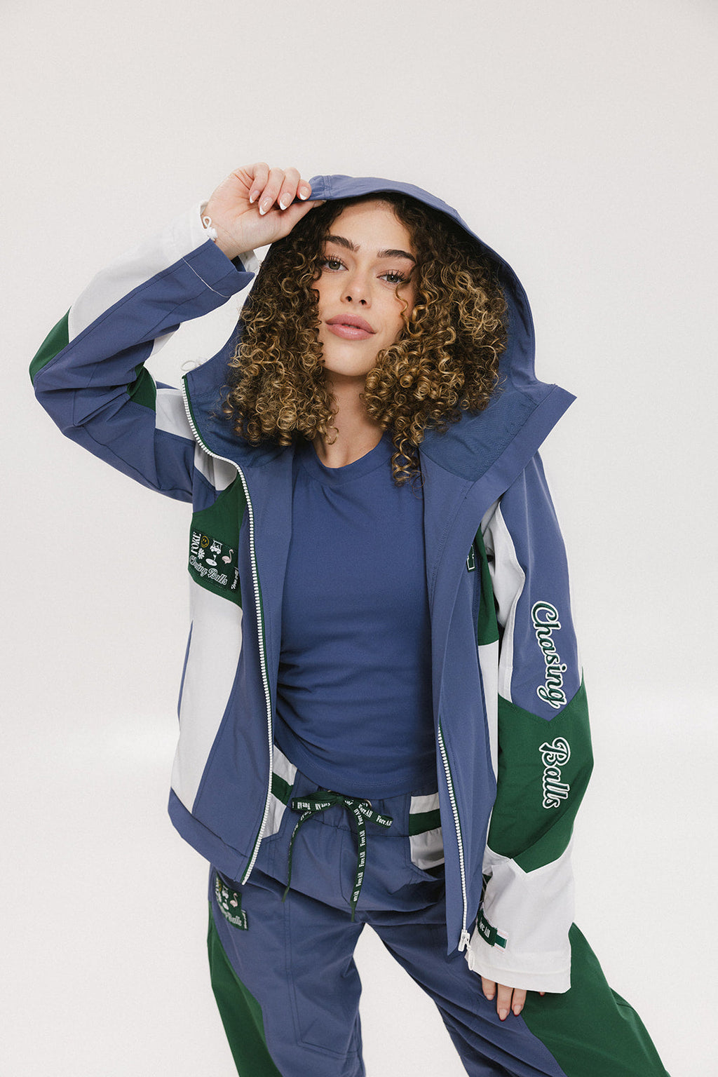 A female model wearing a blue and green color-blocked warm-up jacket with zipper pockets and a high neck collar.