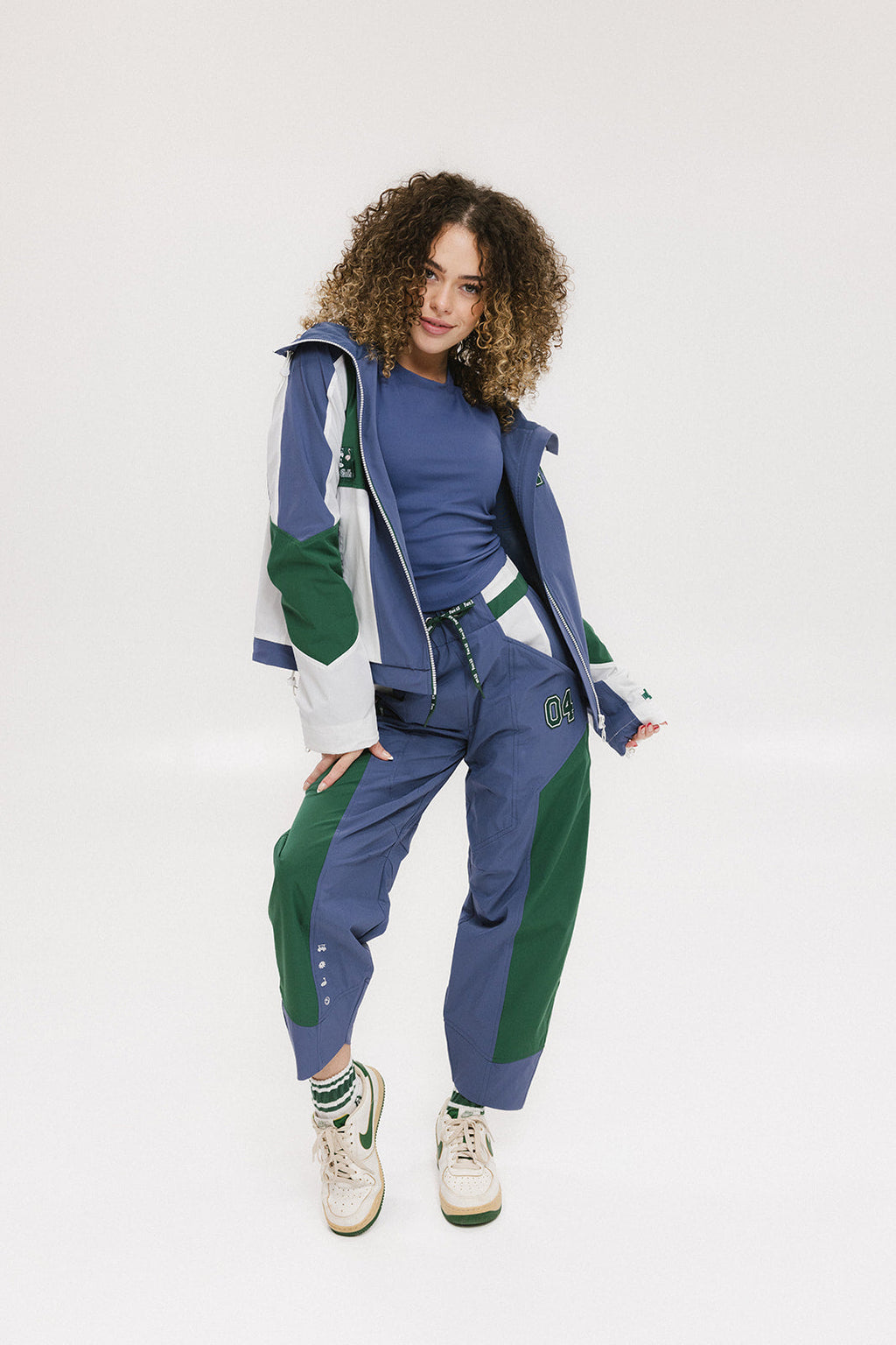A female model wearing a blue and green color-blocked warm-up jacket with zipper pockets and a high neck collar.
