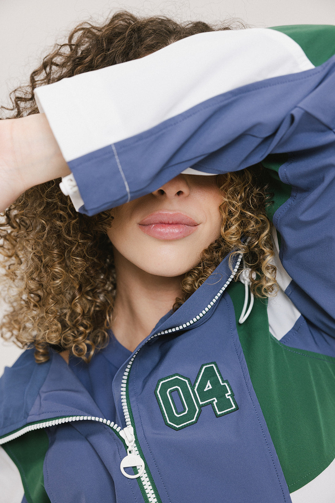 A female model wearing a blue and green color-blocked warm-up jacket with zipper pockets and a high neck collar.