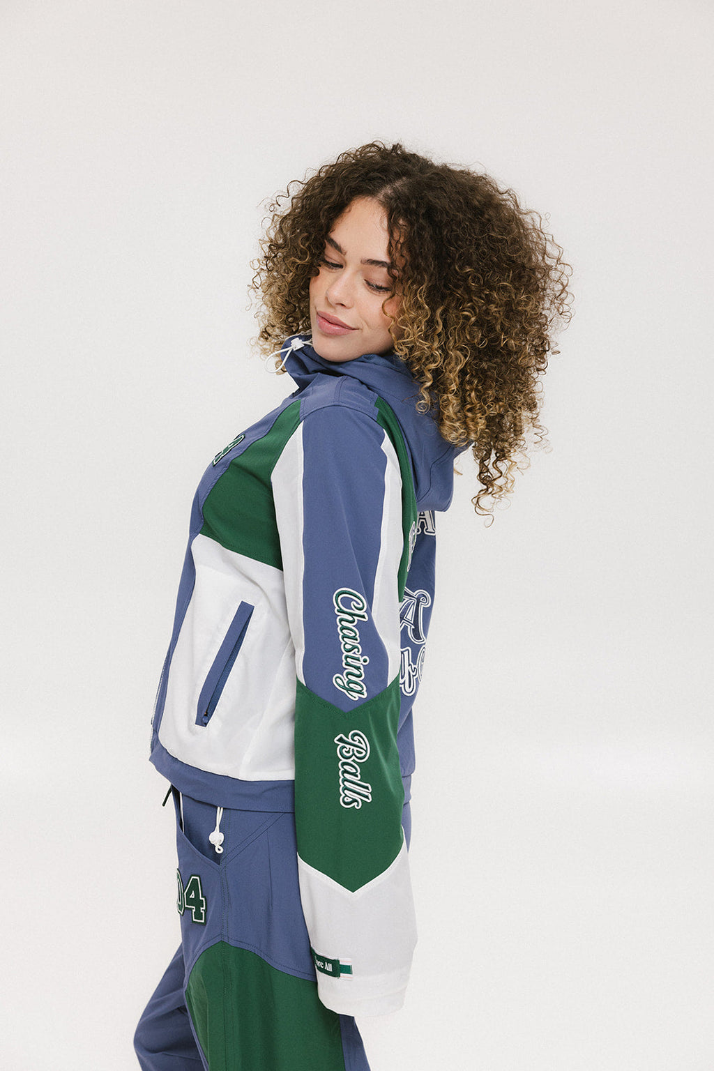 A female model wearing a blue and green color-blocked warm-up jacket with zipper pockets and a high neck collar.
