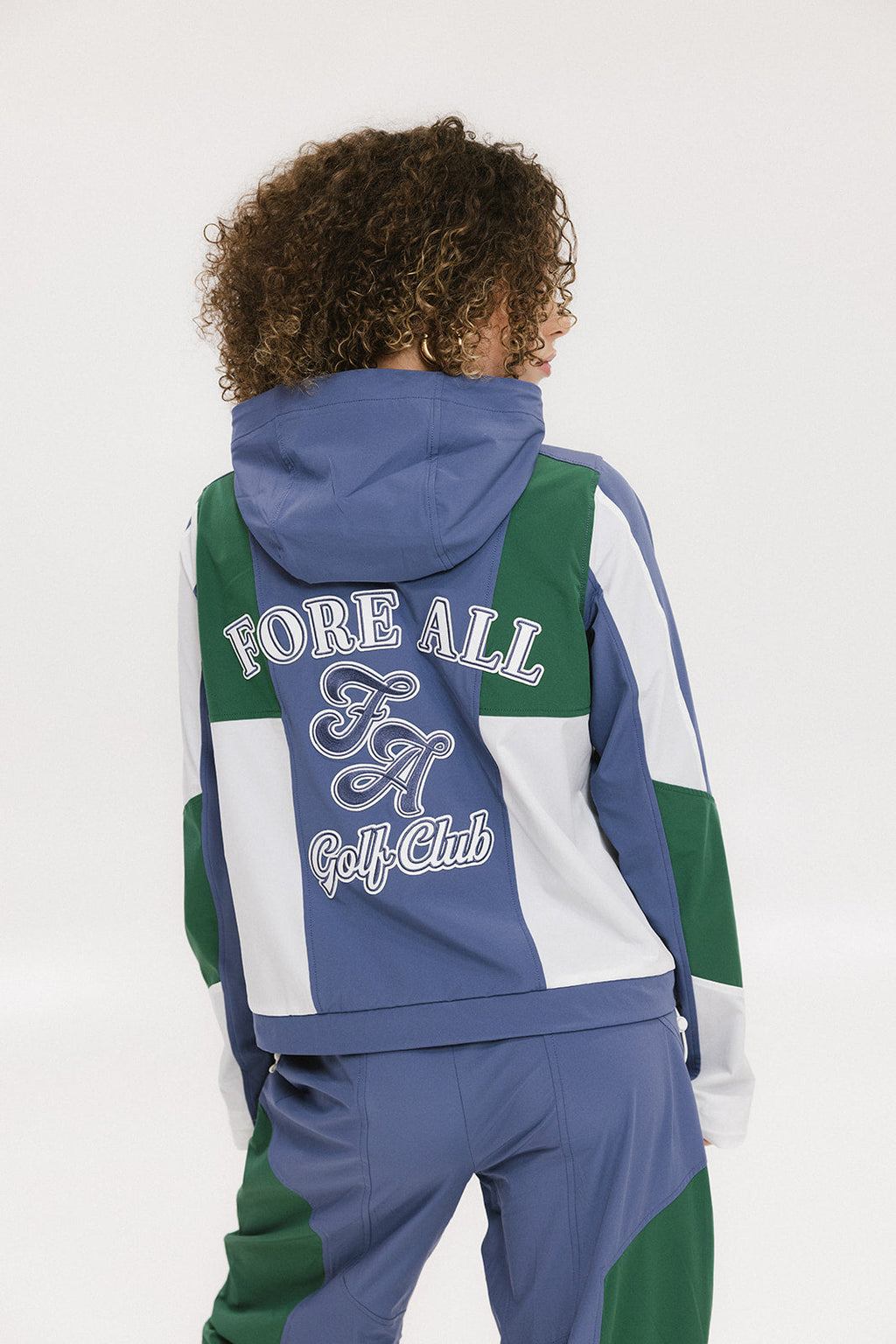 A female model wearing a blue and green color-blocked warm-up jacket with zipper pockets and a high neck collar.