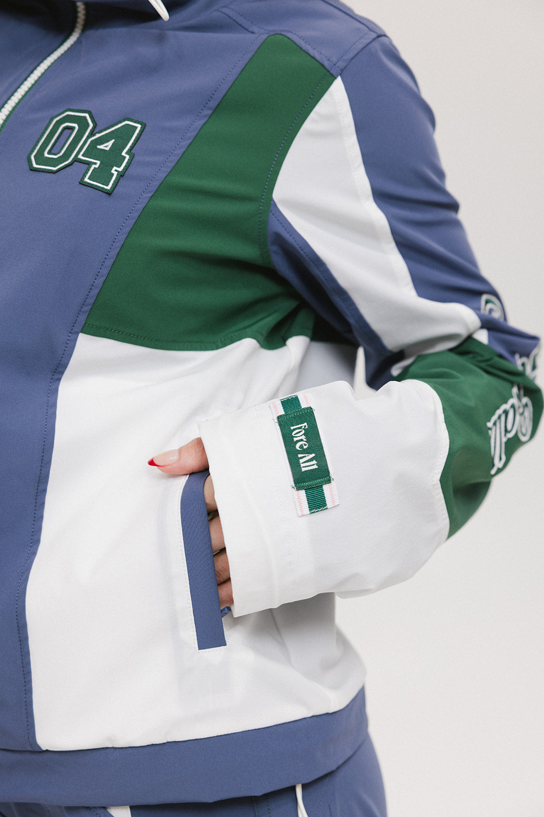A female model wearing a blue and green color-blocked warm-up jacket with zipper pockets and a high neck collar.