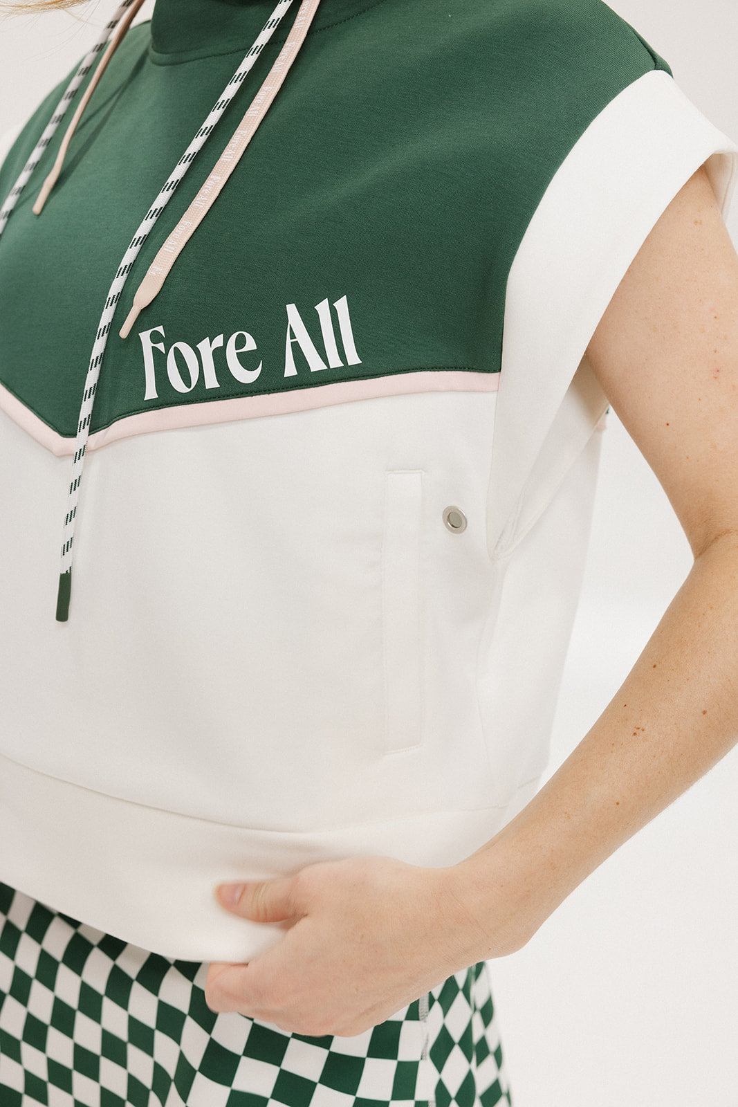 A person wearing a green and white sleeveless hoodie with a hood, matching green and white checkered hat, and green and white checkered skirt. The hoodie has white text and branding on it.