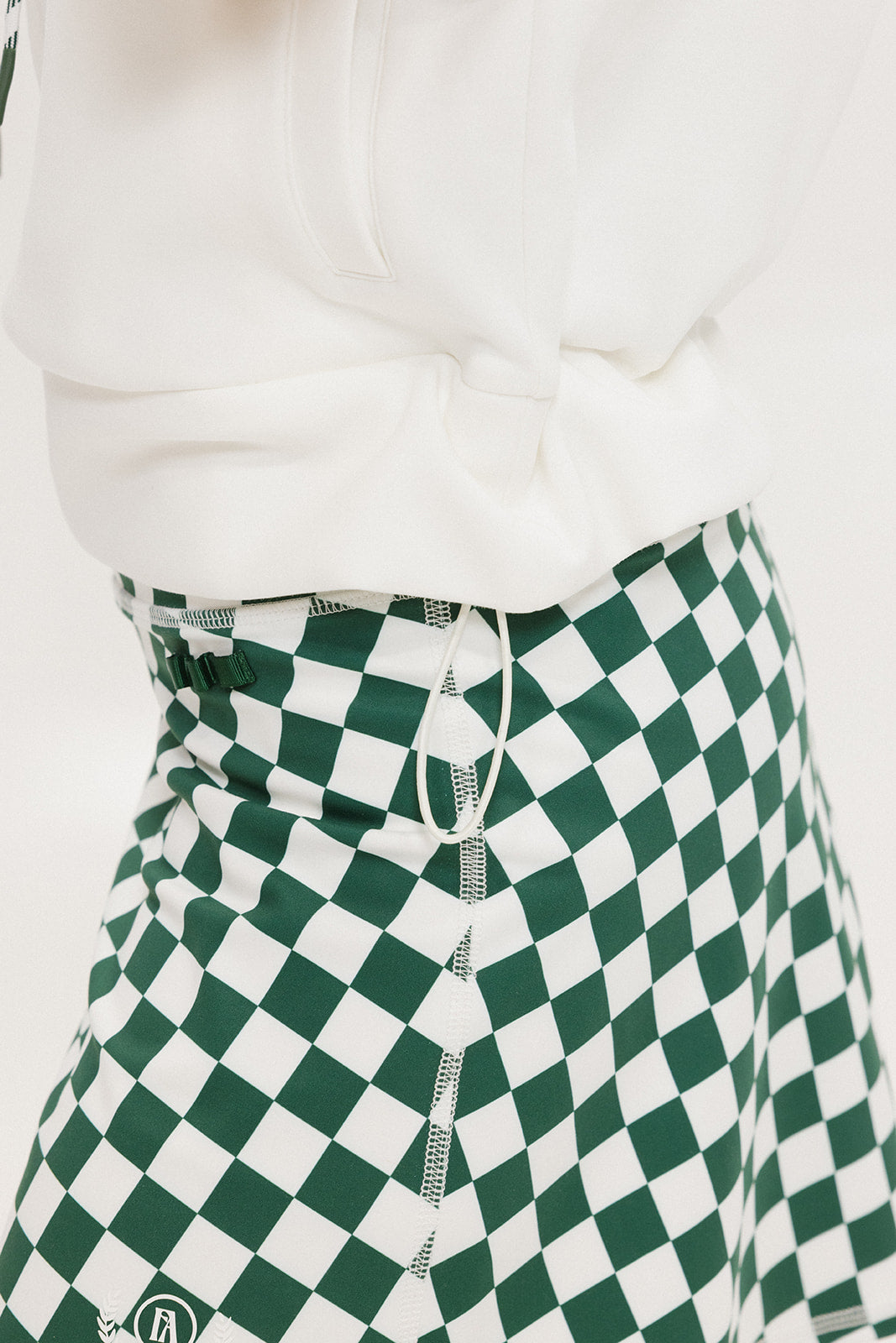 A person wearing a green and white sleeveless hoodie with a hood, matching green and white checkered hat, and green and white checkered skirt. The hoodie has white text and branding on it.