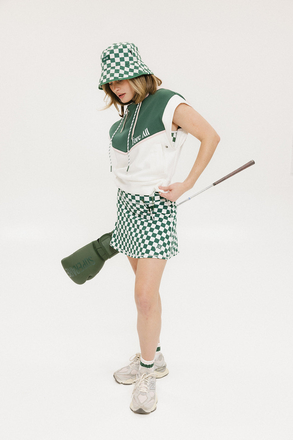 A person wearing a green and white sleeveless hoodie with a hood, matching green and white checkered hat, and green and white checkered skirt. The hoodie has white text and branding on it.