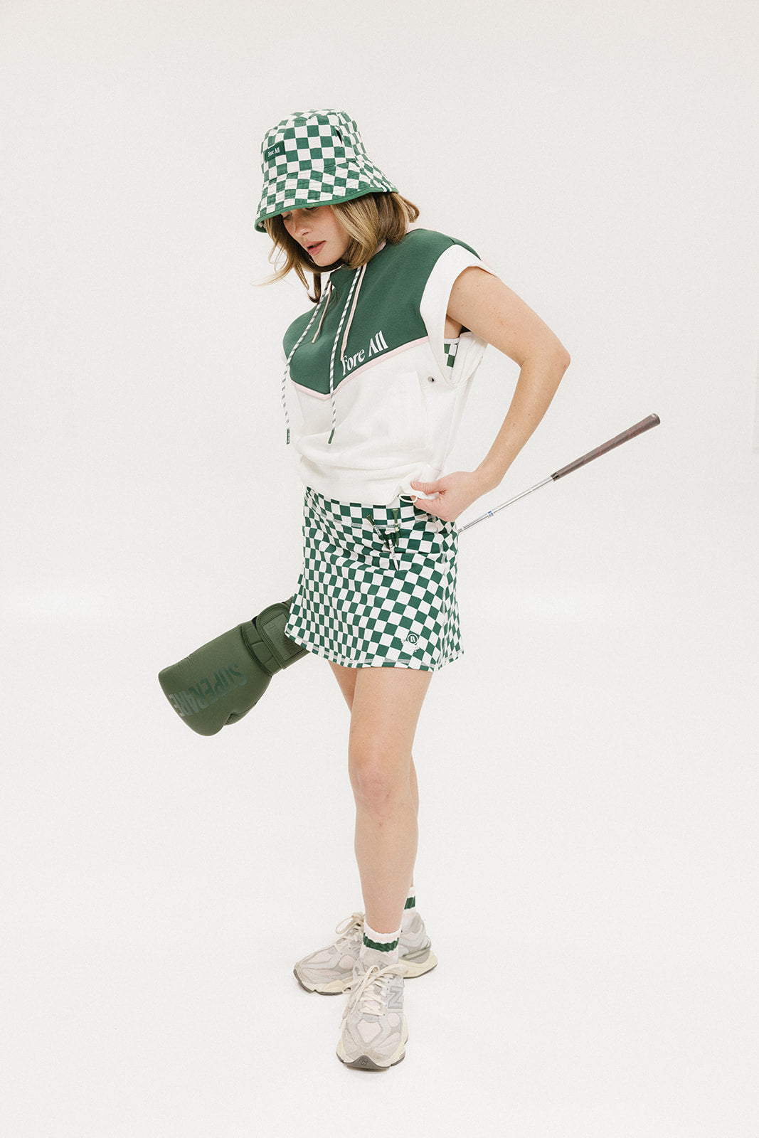 A person wearing a green and white sleeveless hoodie with a hood, matching green and white checkered hat, and green and white checkered skirt. The hoodie has white text and branding on it.