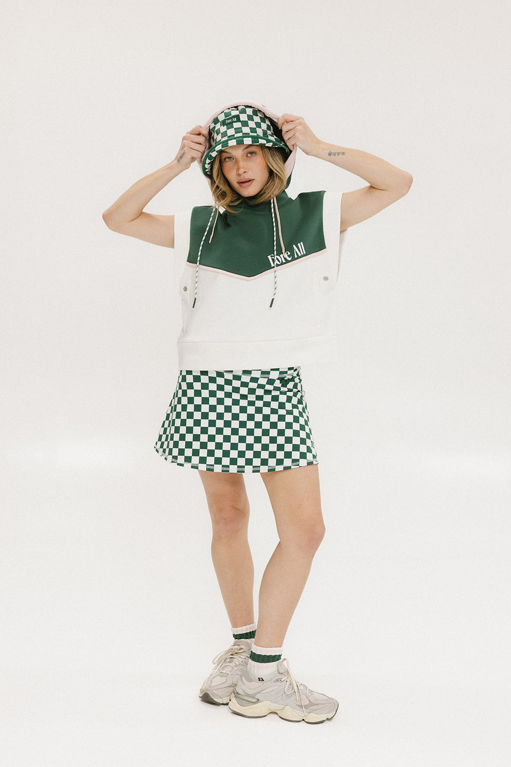A person wearing a green and white sleeveless hoodie with a hood, matching green and white checkered hat, and green and white checkered skirt. The hoodie has white text and branding on it.