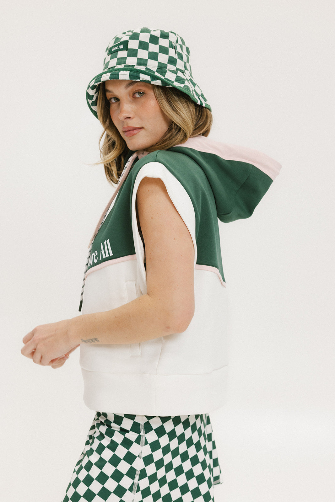 A person wearing a green and white sleeveless hoodie with a hood, matching green and white checkered hat, and green and white checkered skirt. The hoodie has white text and branding on it.