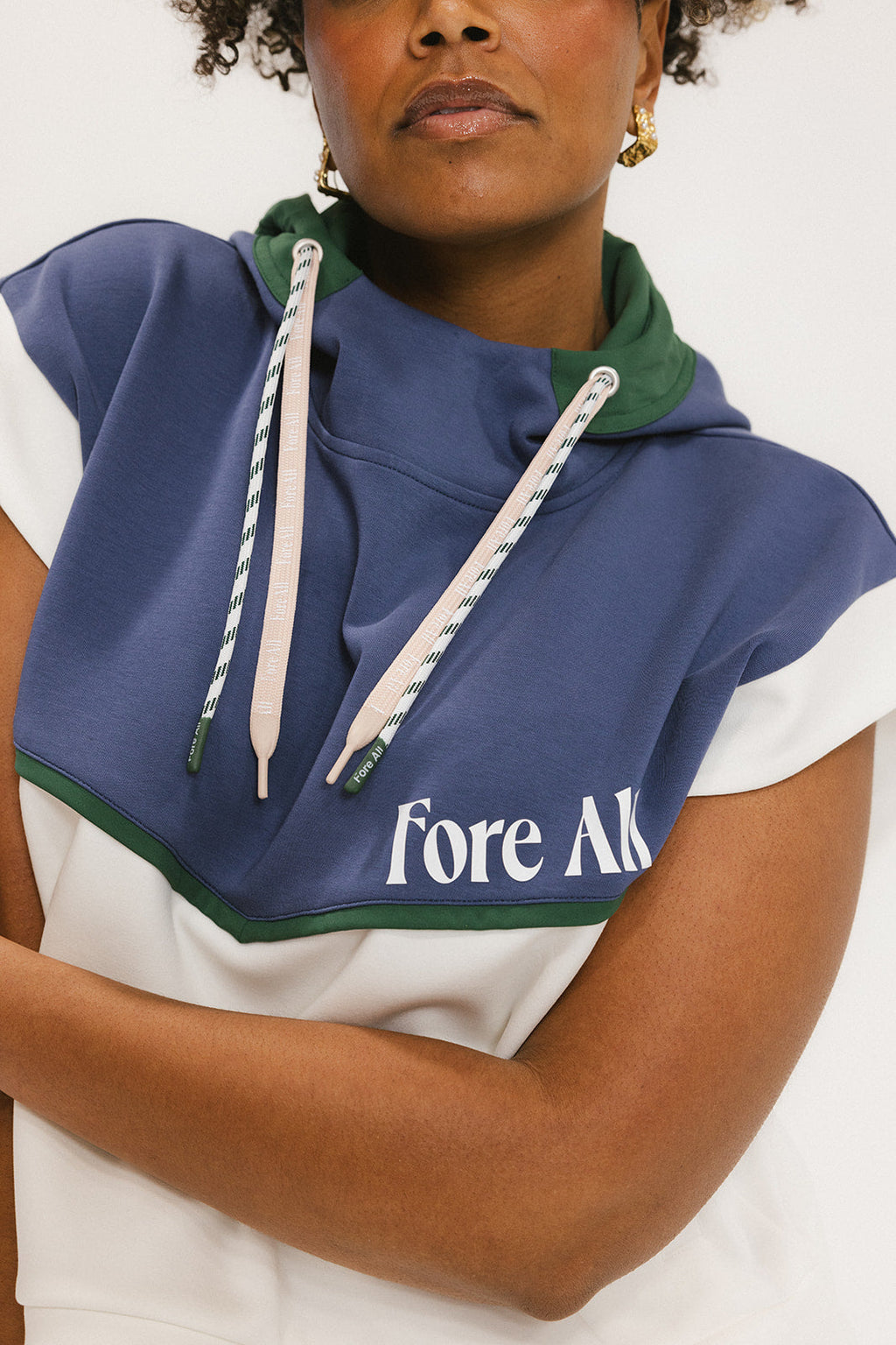 A person wearing a sleeveless hoodie with colorblocking and branding on the chest, in indigo, white, and green colors.