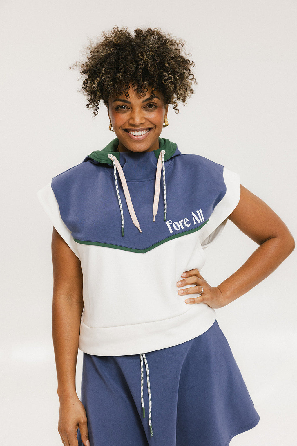 A person wearing a sleeveless hoodie with colorblocking and branding on the chest, in indigo, white, and green colors.