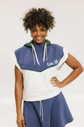 A person wearing a sleeveless hoodie with colorblocking and branding on the chest, in indigo, white, and green colors.