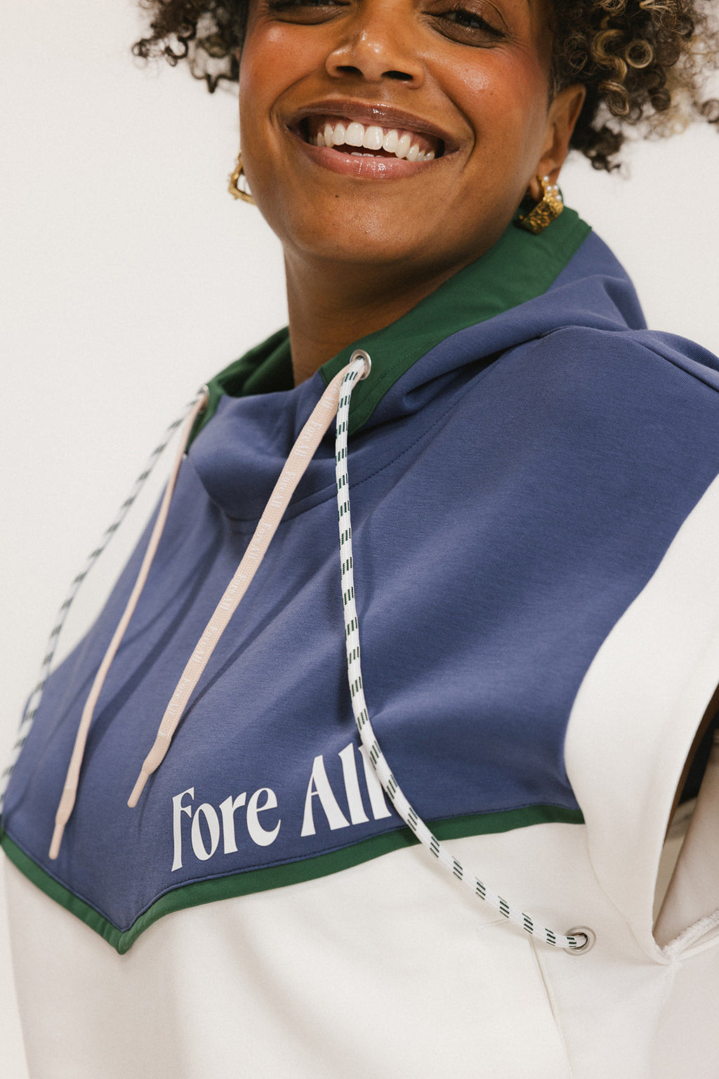 A person wearing a sleeveless hoodie with colorblocking and branding on the chest, in indigo, white, and green colors.