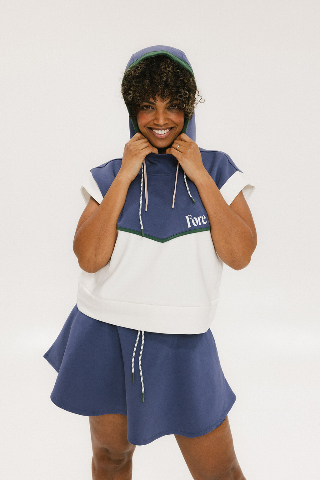 A person wearing a sleeveless hoodie with colorblocking and branding on the chest, in indigo, white, and green colors.