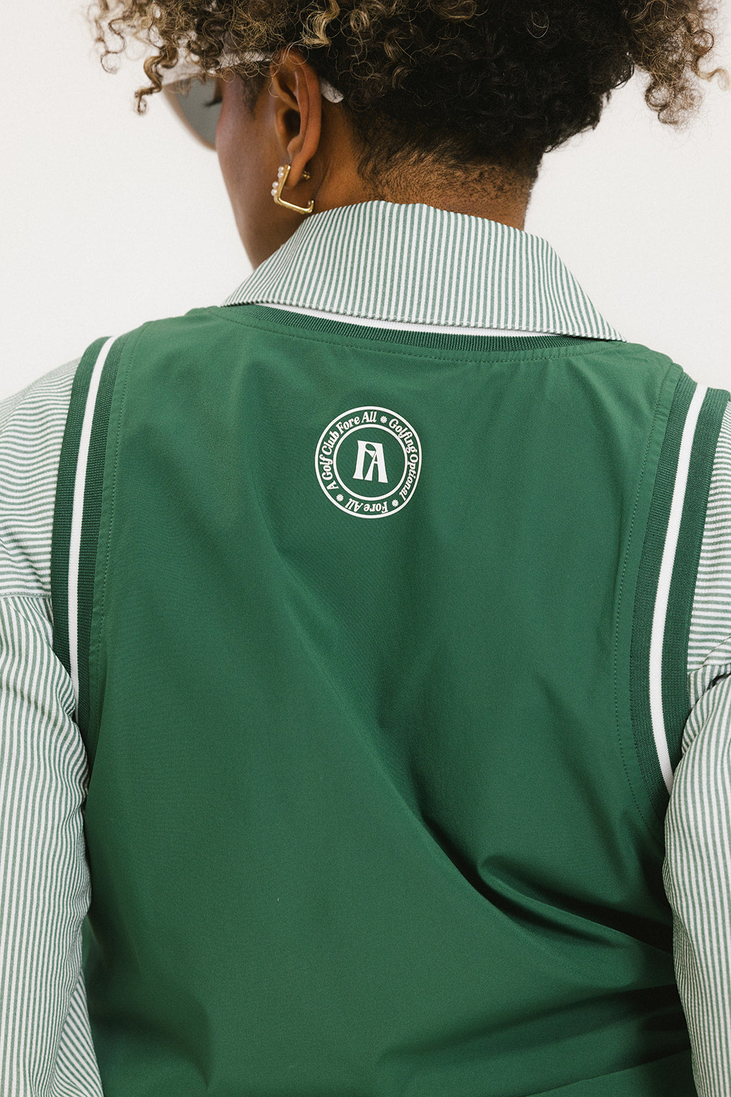 A woman wearing a green sleeveless windbreaker vest with a collar, featuring a white appliquéd logo on the left chest and silicone logos on the upper back.