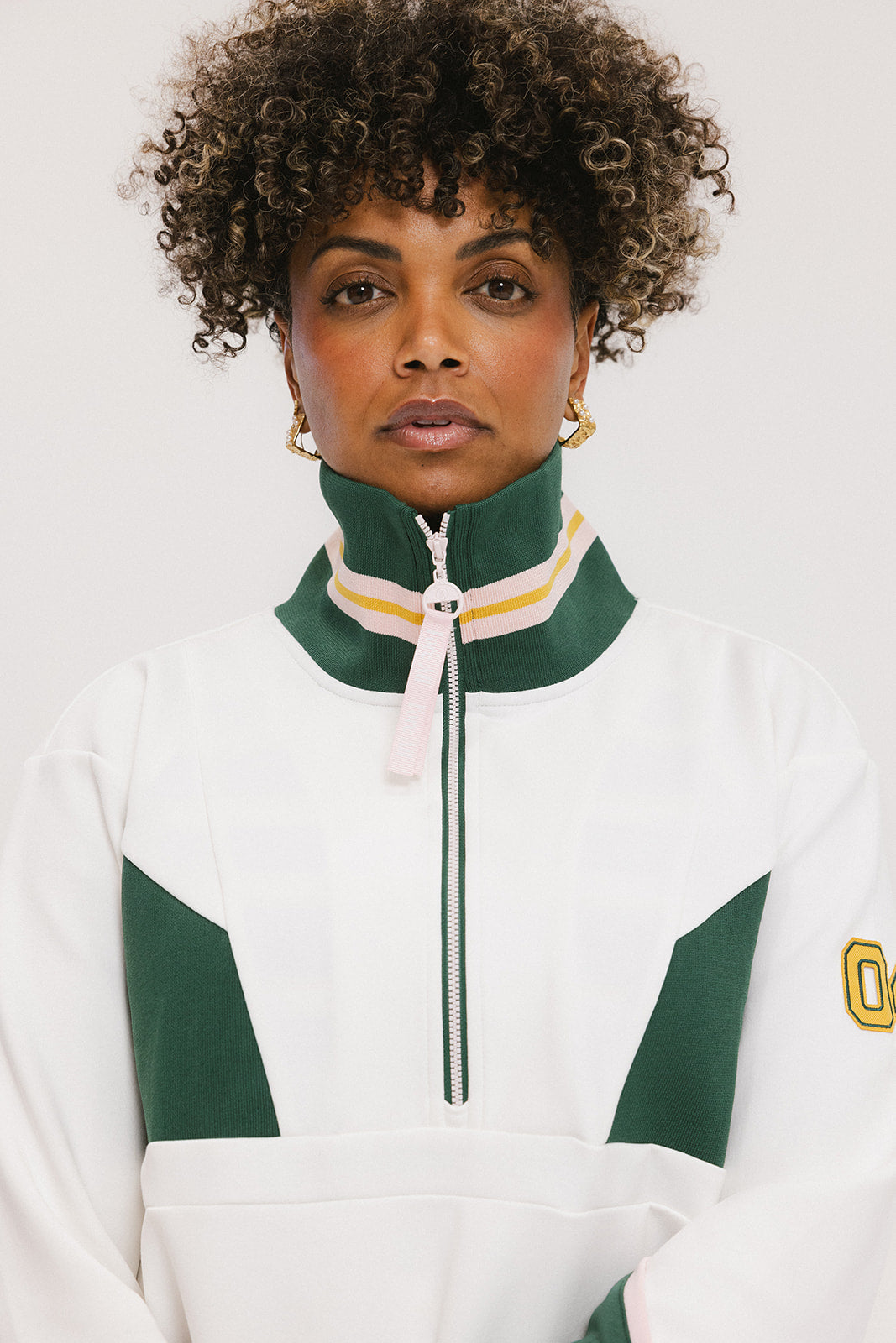 A woman wearing a white half zip sports top with green color blocking and ribbed detailing around the collar, cuffs, and hem. The top features half sleeve design with a signature logo on the chest.