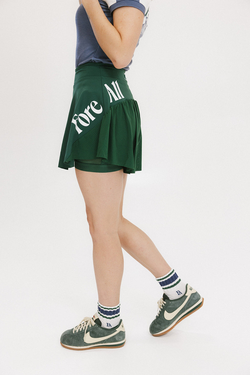 A person wearing a green skirt with a mesh back, paired with a blue top and green sneakers. The skirt has a high waist and a mid-thigh length cut.