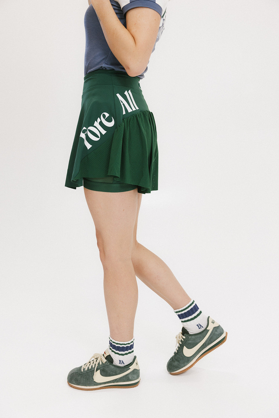 A person wearing a green skirt with a mesh back, paired with a blue top and green sneakers. The skirt has a high waist and a mid-thigh length cut.